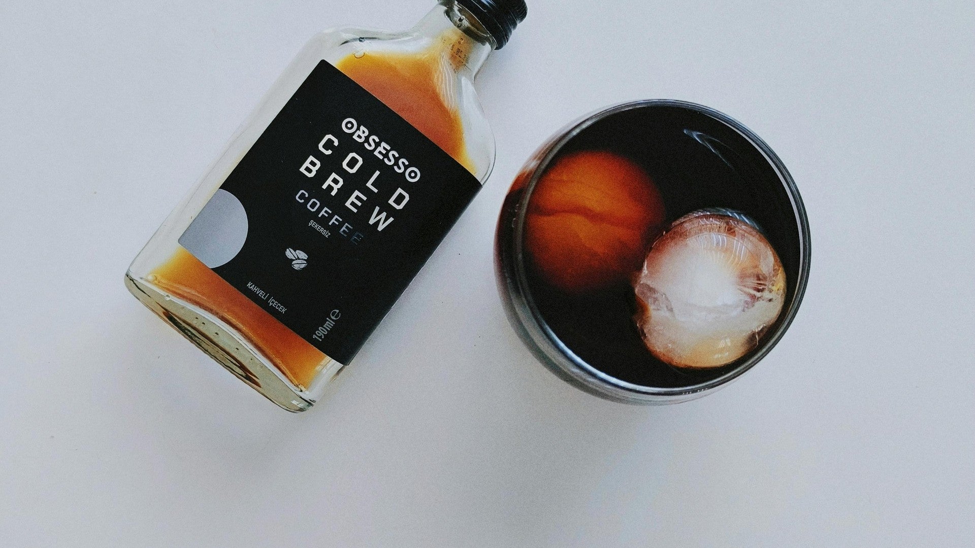 Cold brew coffee bottle with large ice sphere in glass — flatlay on white background
